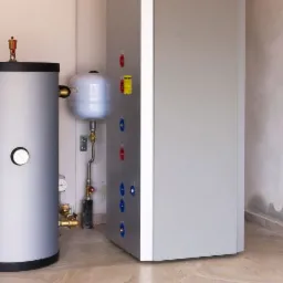 Szary zasobnik ciepłej wody użytkowej z manometrem i rurami w izolacji, obok biały zbiornik wyrównawczy i skrzynka sterownicza z przyłączami w kotłowni z beżową podłogą.