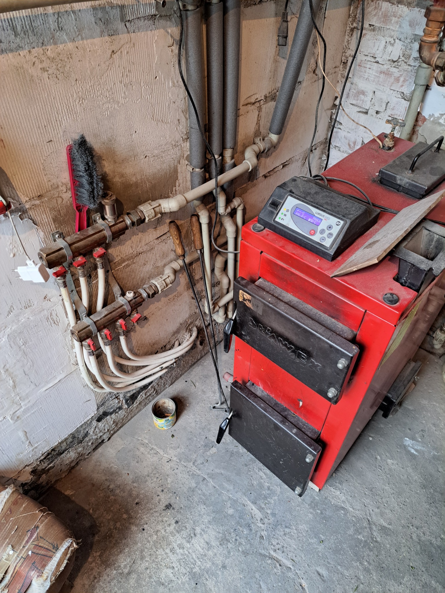 Czerwony piec na paliwo stałe marki Drawex z czarnymi drzwiczkami, zamontowany w piwnicy z widocznymi rurami, zaworami i narzędziami do obsługi pieca.