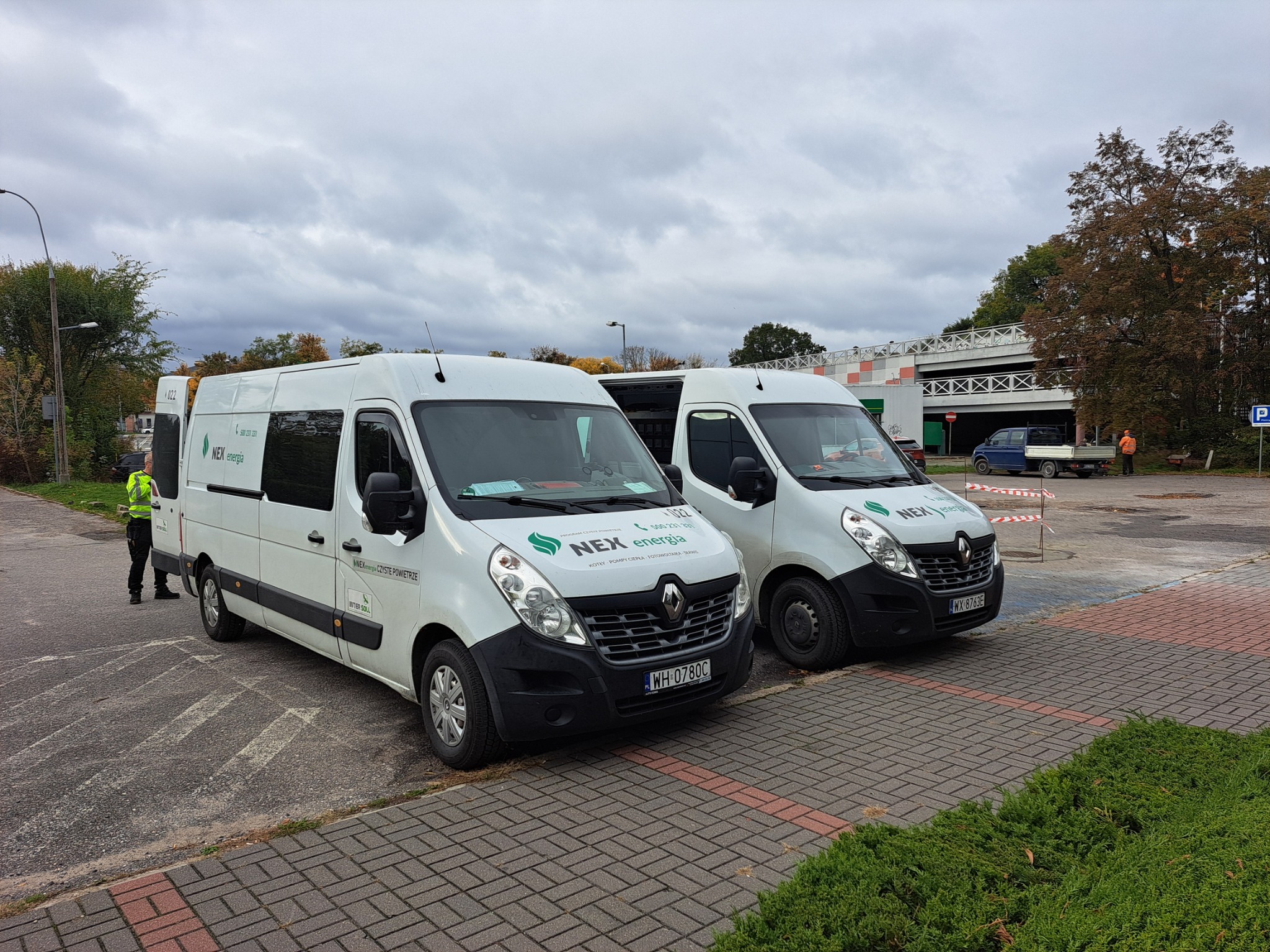 Dwa białe samochody dostawcze firmy NEX energia z logo Czyste Powietrze, kotły, pompy ciepła, fotowoltaika, serwis, zaparkowane na brukowanym placu w pochmurny dzień.