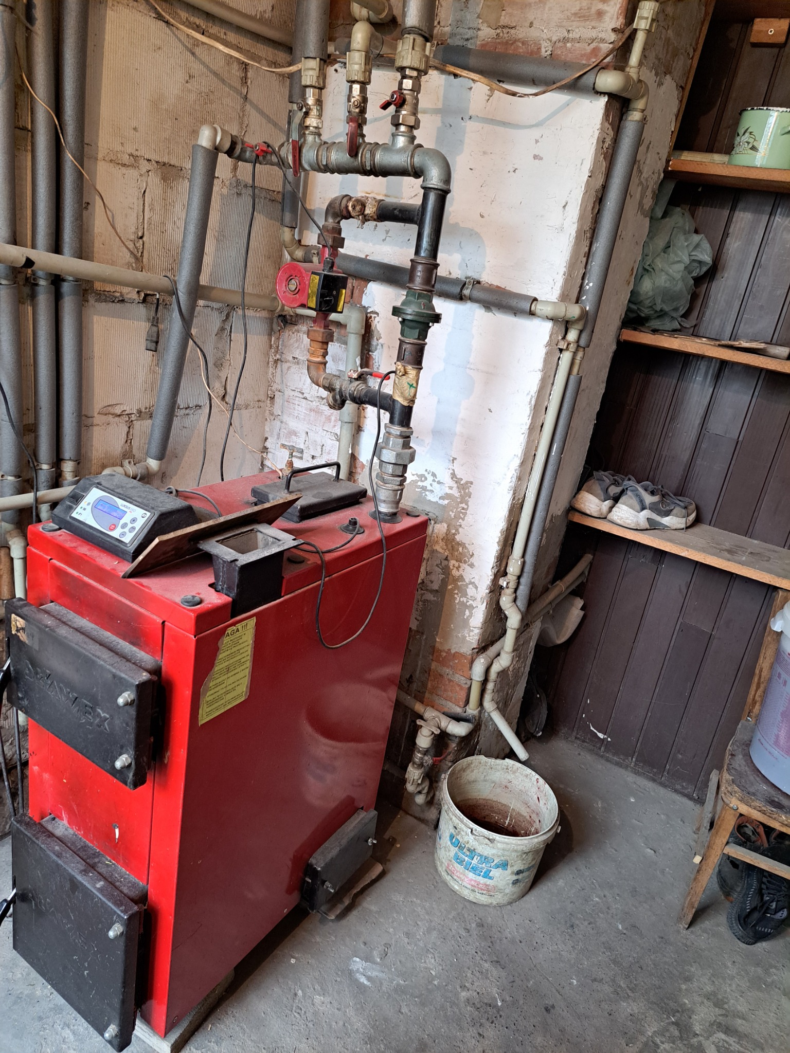 Czerwony piec z czarnymi drzwiczkami i elektronicznym sterownikiem w kotłowni, widoczne rury instalacji centralnego ogrzewania i fragment regału z butami.