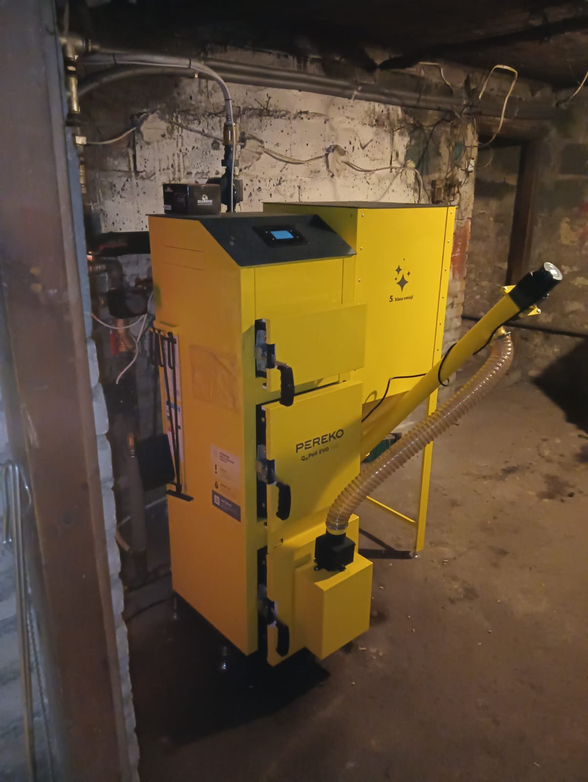 Żółty piec na pellet marki PEREKO Q-Pell EVO 30 w kotłowni o ścianach z widoczną cegłą i instalacjami.