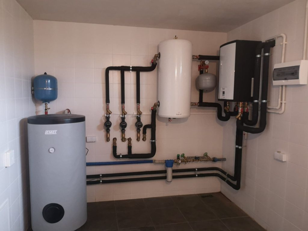 Kotłownia z dwoma zbiornikami akumulacyjnymi, pompą ciepła, rozdzielaczami z zaworami, filtrem wody i instalacją hydrauliczną, całość w pomieszczeniu wyłożonym białymi płytkami.