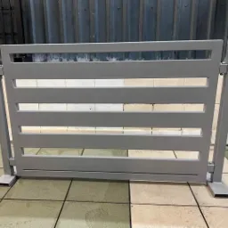 Szare, poziome przęsło ogrodzeniowe z prostokątnymi szczeblami, umieszczone na podłodze z płytek. Widoczne spawane łączenia i metalowe podstawy słupków.