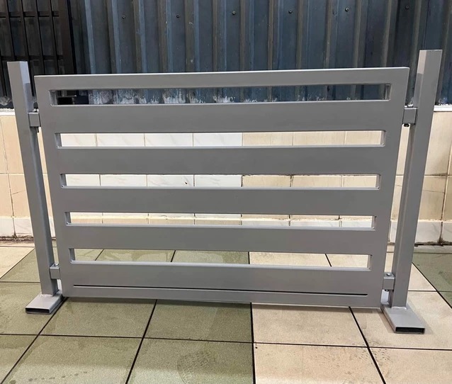 Szare, poziome przęsło ogrodzeniowe z prostokątnymi szczeblami, umieszczone na podłodze z płytek. Widoczne spawane łączenia i metalowe podstawy słupków.