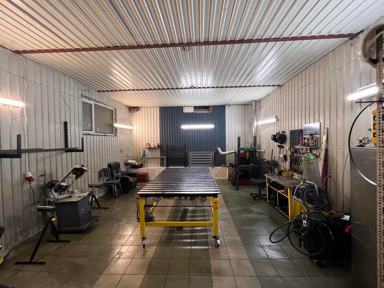 Warsztat z żółtym stołem spawalniczym na kółkach, wyposażony w narzędzia i materiały, oświetlony świetlówkami, z blachą falistą na ścianach i suficie.