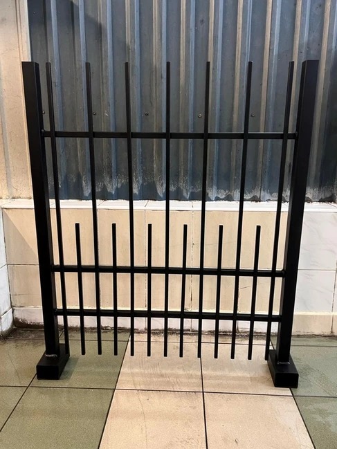 Czarne, metalowe przęsło ogrodzeniowe z pionowymi szczeblami o zróżnicowanej długości, ustawione na podłodze w pomieszczeniu z blachą falistą w tle.