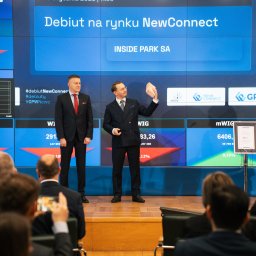 dabrowskistudio.pl Arkadiusz Dąbrowski - Dwóch mężczyzn w garniturach na scenie podczas debiutu firmy na rynku NewConnect, trzymający cegłę na tle ekranów z danymi giełdowymi i logo firmy.
