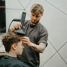 dabrowskistudio.pl Arkadiusz Dąbrowski - Fryzjer stylizuje kręcone włosy klienta za pomocą dyfuzora, widoczne tatuaże na rękach i szyi fryzjera, minimalistyczne wnętrze salonu.