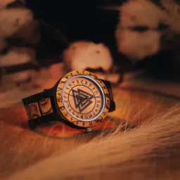 Drewniany zegarek z runami i symbolem Walknut na tarczy, leżący na drewnianej powierzchni z dekoracyjnymi trawami i gałązkami bawełny w tle.