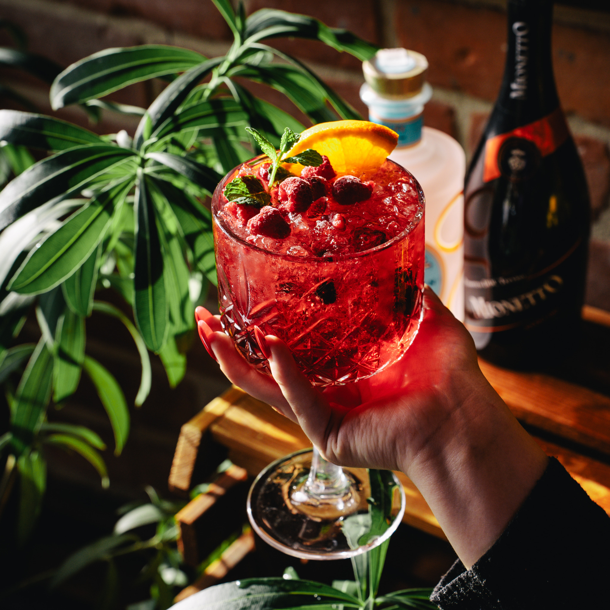 Kryształowy kieliszek wypełniony czerwonym koktajlem z lodem, malinami, pomarańczą i miętą, trzymany przez osobę z czerwonymi paznokciami na tle ceglanej ściany, butelki wina musującego i zielonej...