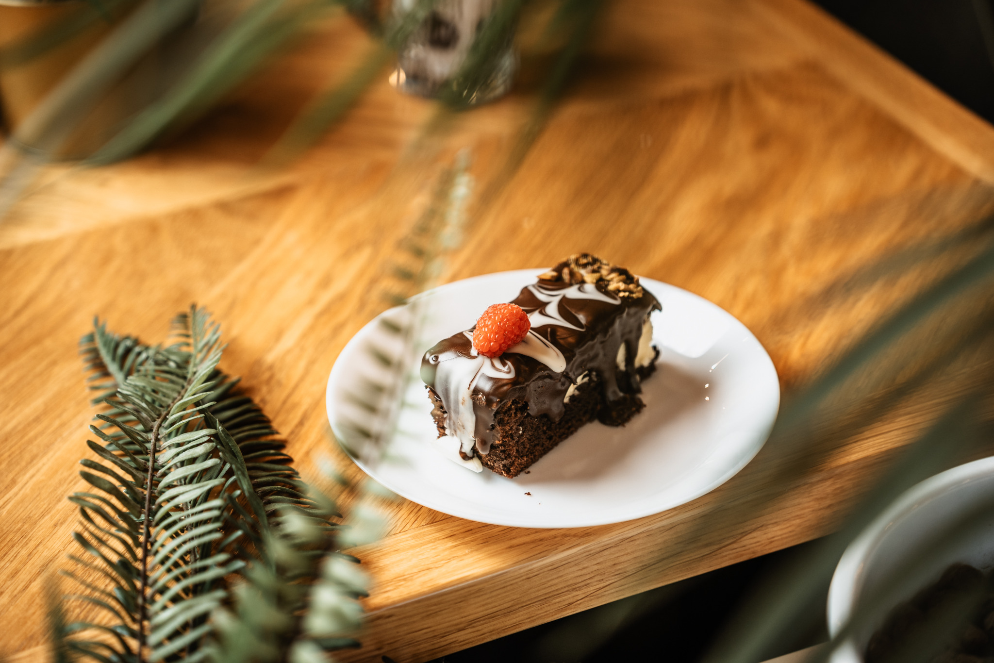 Kawałek czekoladowego ciasta z polewą i maliną na białym talerzu, umieszczony na drewnianym blacie z dekoracją z gałązek.