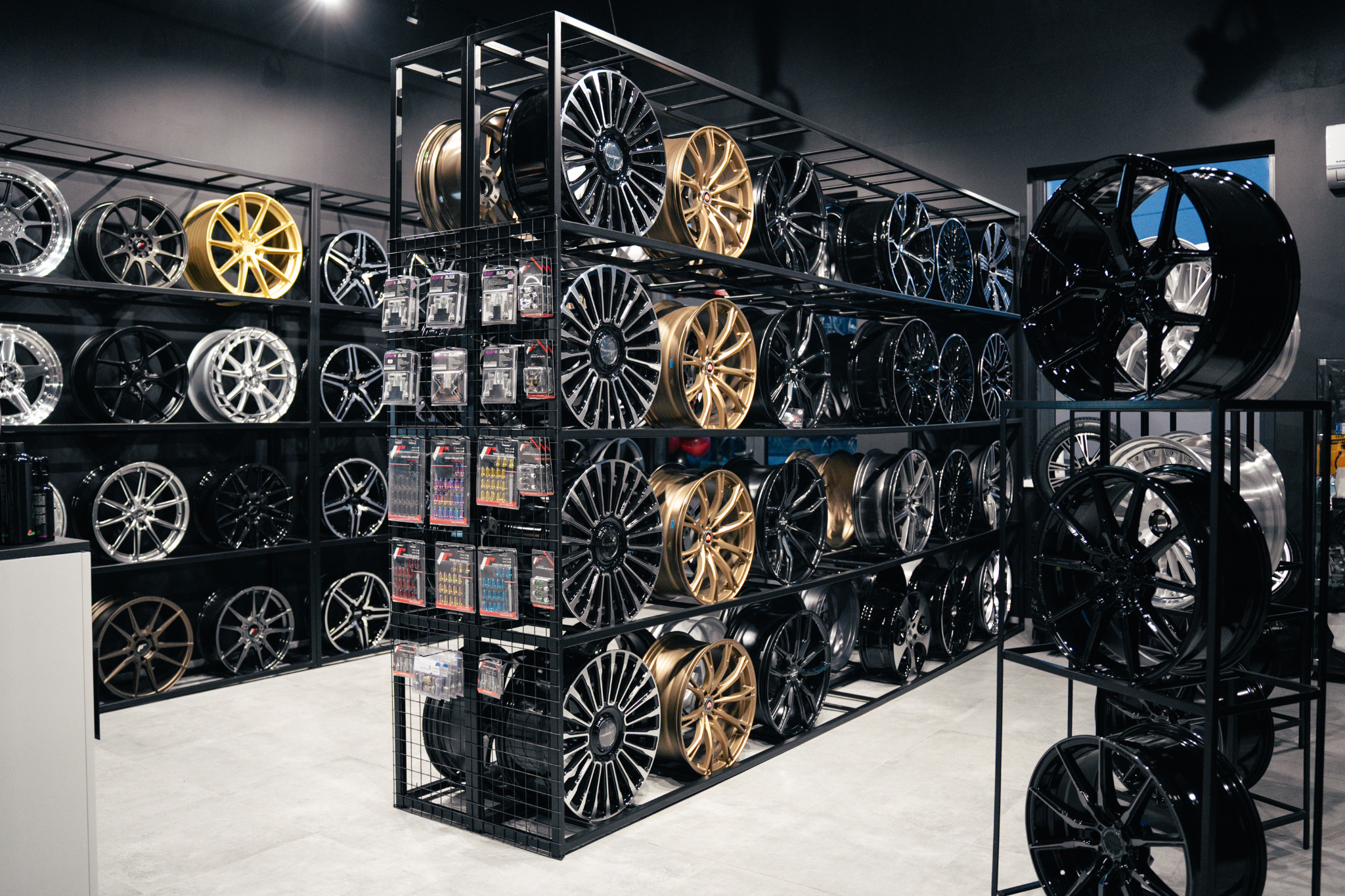 Ekspozycja felg aluminiowych różnych wzorów i kolorów (złote, czarne, srebrne) na metalowym regale w sklepie, widoczne także akcesoria montażowe.