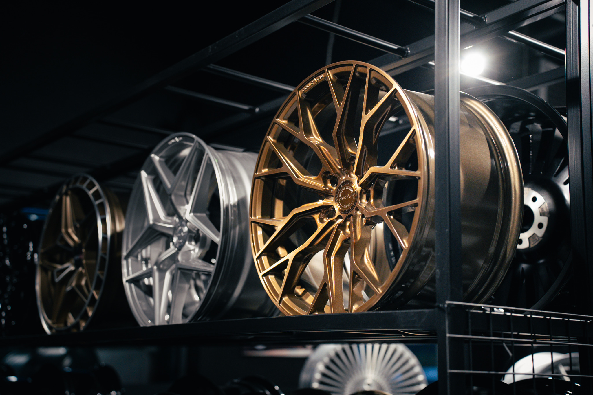 Rząd felg aluminiowych w odcieniach złota, srebra i czerni, umieszczonych na metalowym regale w warsztacie, z widocznymi detalami szprych i wykończenia.