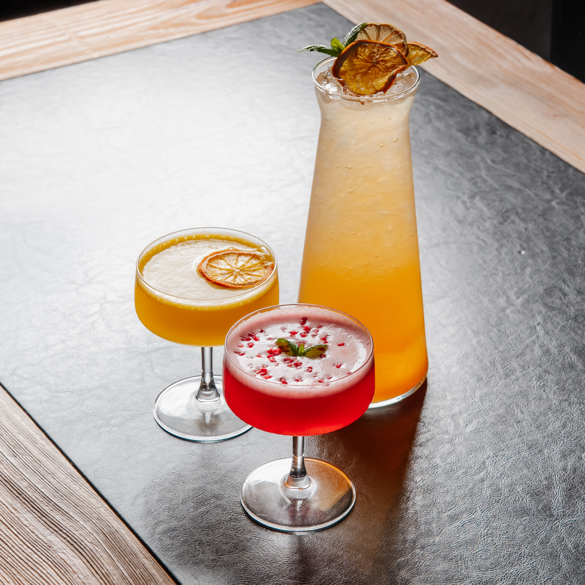 Trzy kolorowe koktajle w szklanych kieliszkach, udekorowane suszonymi plastrami pomarańczy i miętą, stoją na czarnym blacie z drewnianą ramą.