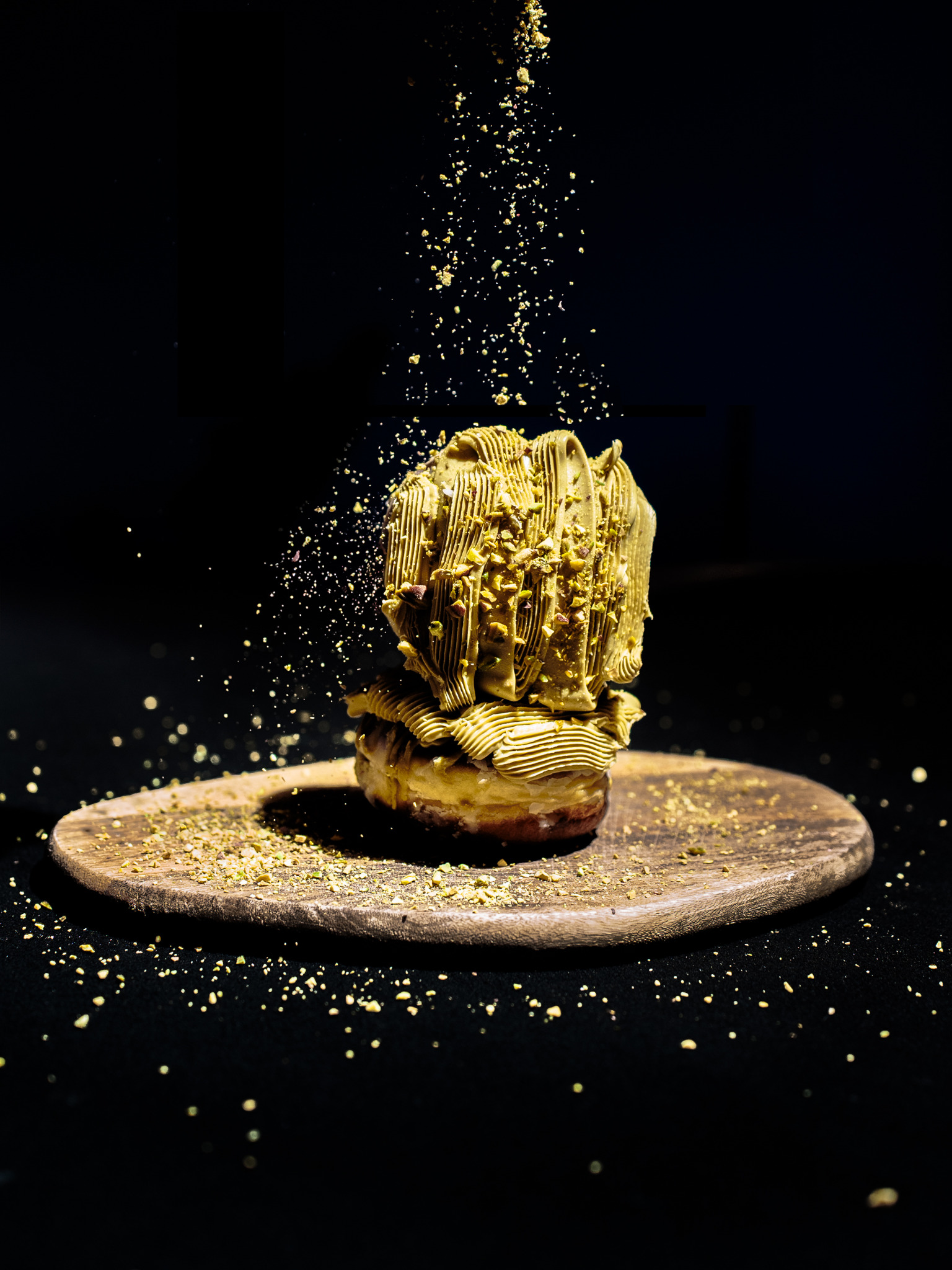 Donut z kremem pistacjowym i posypką, prezentowany na drewnianej desce na czarnym tle, z opadającymi drobinkami posypki.