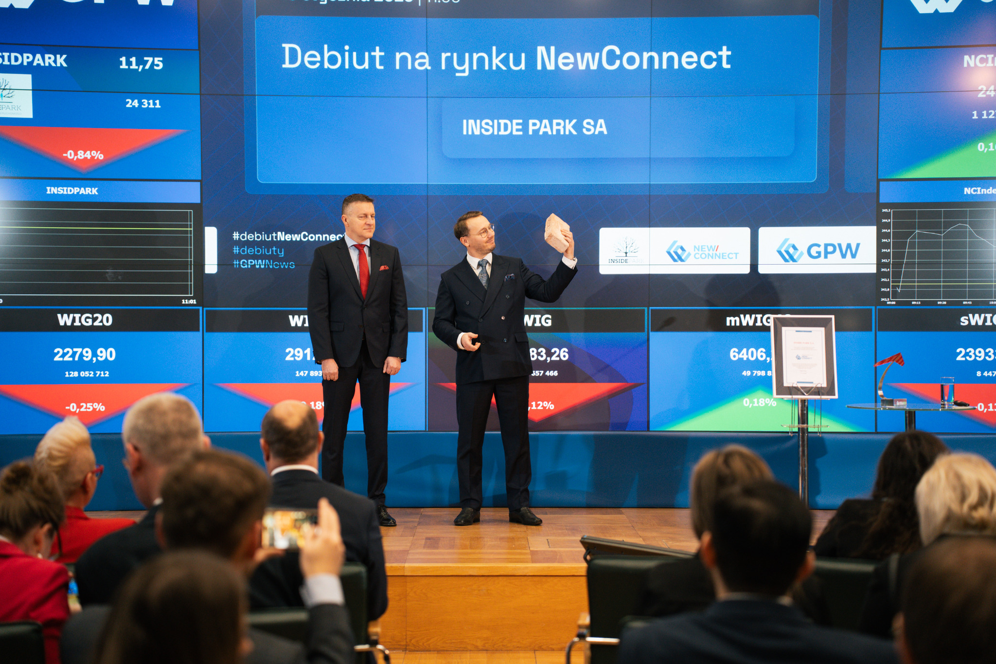 Dwóch mężczyzn w garniturach na scenie podczas debiutu firmy na rynku NewConnect, trzymający cegłę na tle ekranów z danymi giełdowymi i logo firmy.