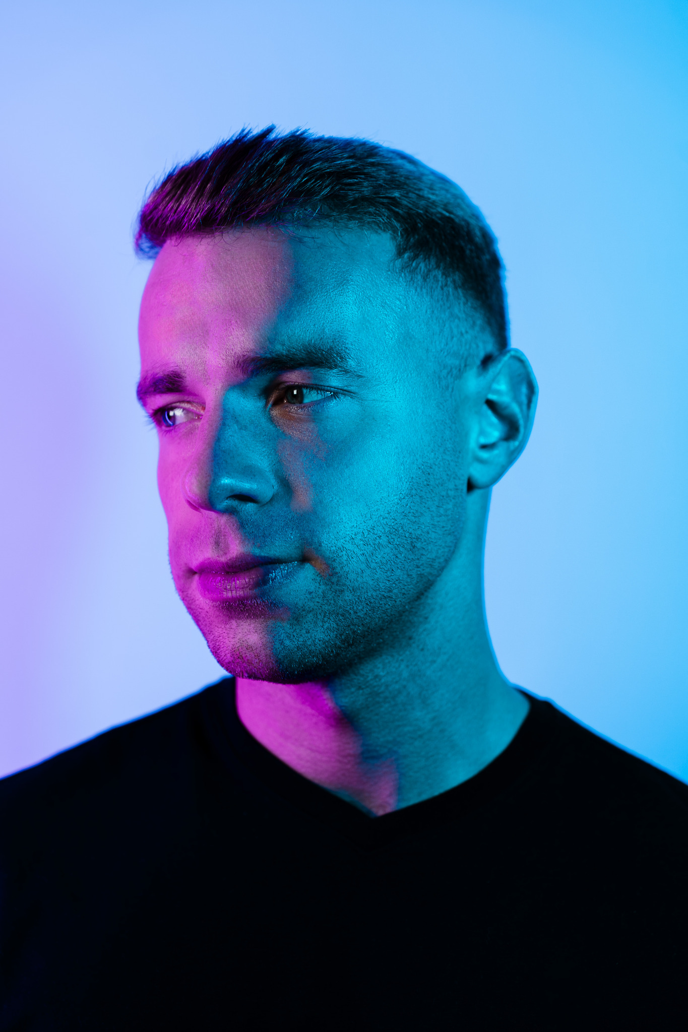 Portret mężczyzny z krótkimi włosami, oświetlonego niebiesko-różowym światłem, spoglądającego w lewo, ubrany w czarny T-shirt.