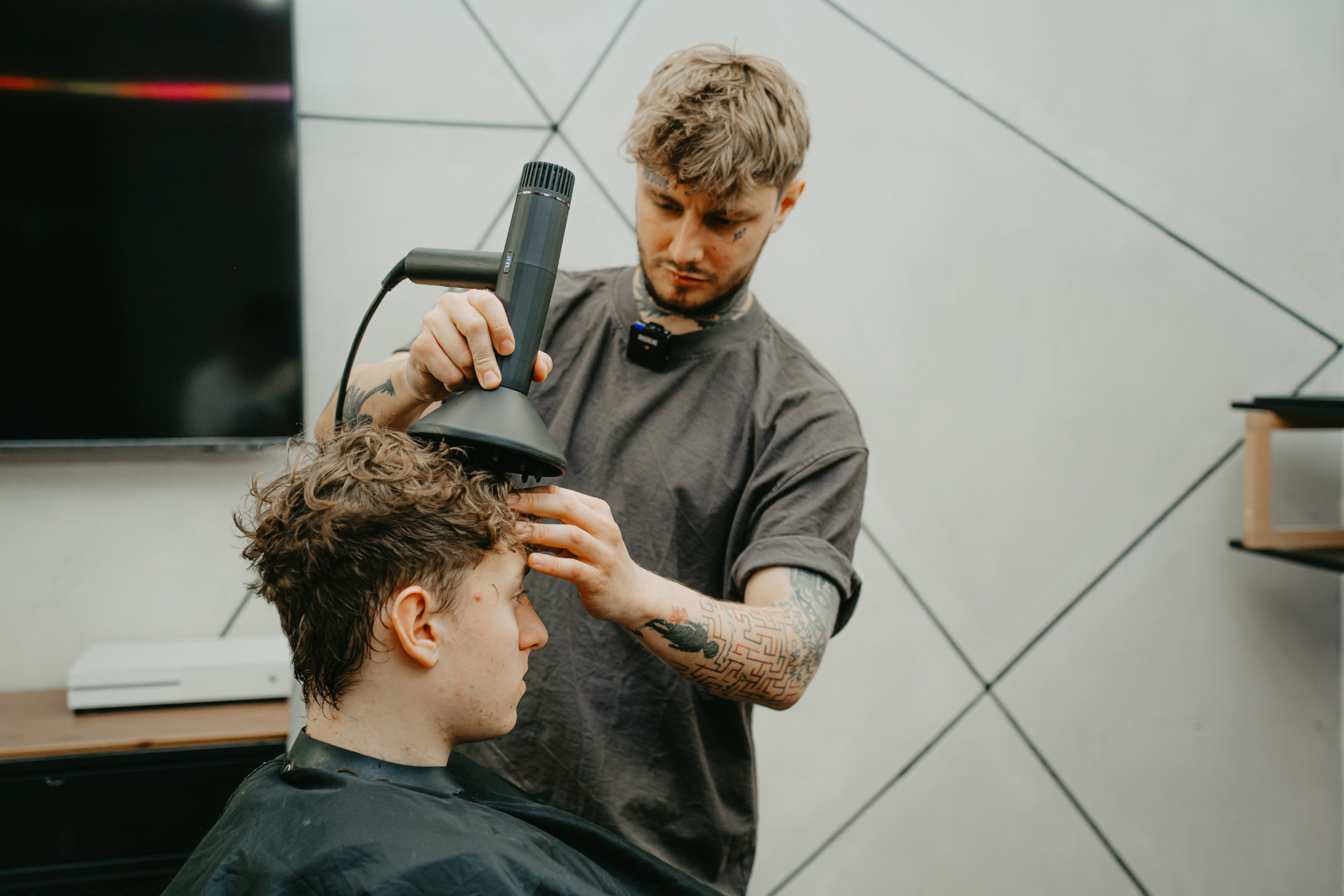 Fryzjer stylizuje kręcone włosy klienta za pomocą dyfuzora, widoczne tatuaże na rękach i szyi fryzjera, minimalistyczne wnętrze salonu.