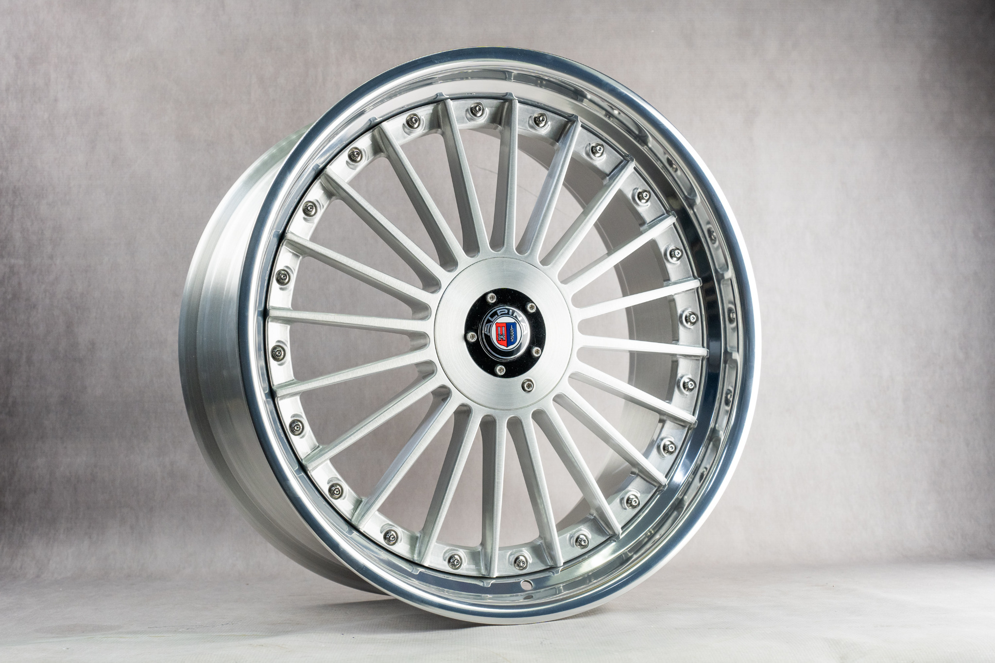 Aluminiowa felga samochodowa z wieloma szprychami, srebrnym wykończeniem i centralnym logo na szarym tle.