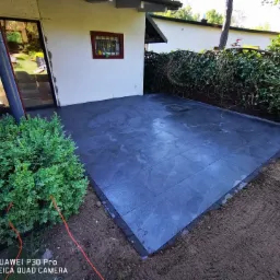 Wykończony taras z ciemnych, prostokątnych płyt, obrzeża z kostki, widok na ogród i budynek z oknami.