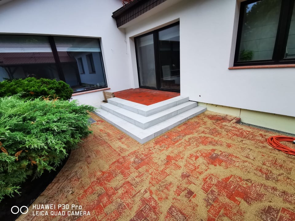 Czerwona kostka brukowa zasypana piaskiem, prowadząca do schodów z białych płyt przed wejściem do budynku z dużymi oknami, obok zielony krzew.