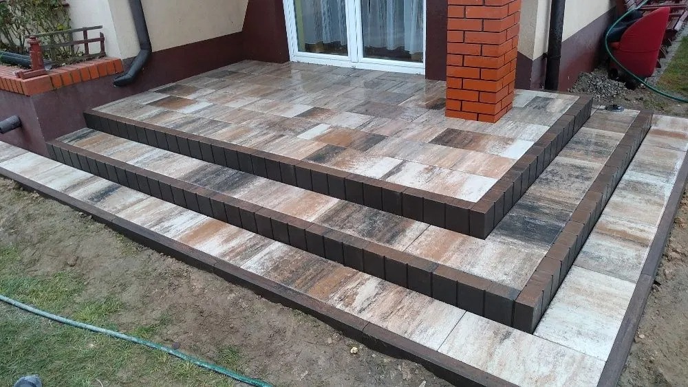Trzy stopnie wejściowe do domu wyłożone kostką brukową o zróżnicowanej kolorystyce, obrzeża wykończone ciemnobrązową kostką. Widoczny fragment elewacji budynku z oknem i ceglanym filarem.