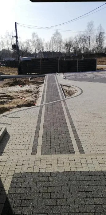 Ułożona ścieżka z kostki brukowej w dwóch odcieniach szarości, prowadząca do domu; widoczne obrzeża i przygotowane podłoże pod dalsze prace brukarskie.