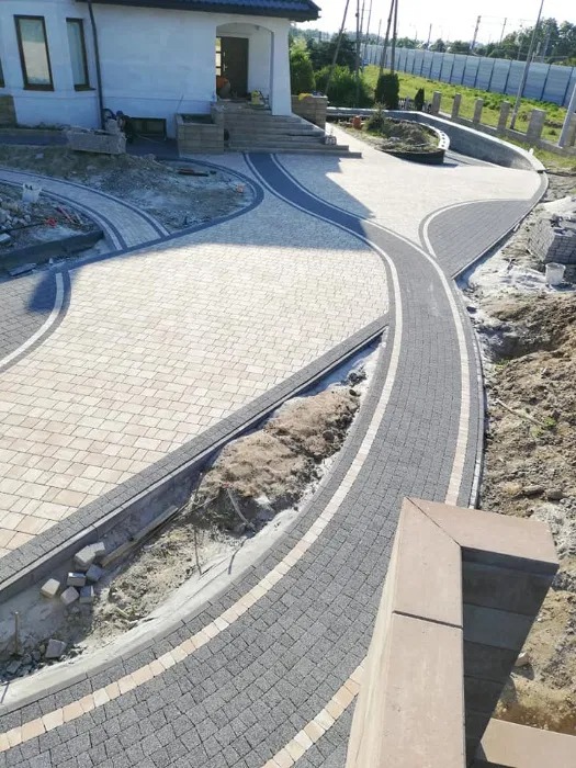 Kaskadowa aranżacja podjazdu z kostki brukowej w odcieniach szarości i beżu, z elementami obrzeży i rabatami ziemnymi, prowadząca do domu z białymi ścianami i schodami.