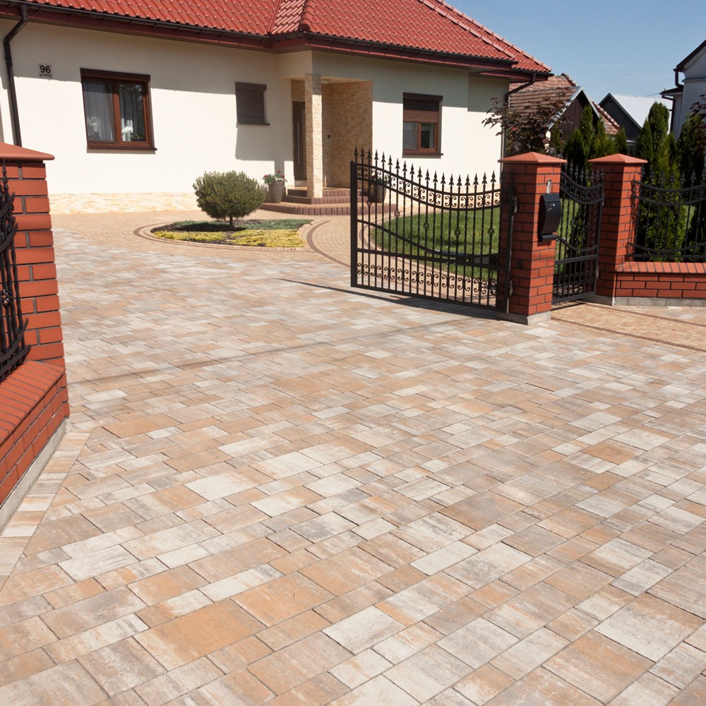 Podjazd z beżowo-szarej kostki brukowej prowadzący do domu z czerwoną dachówką w Gdyni, widoczna brama z czarnego metalu i ceglane słupki.