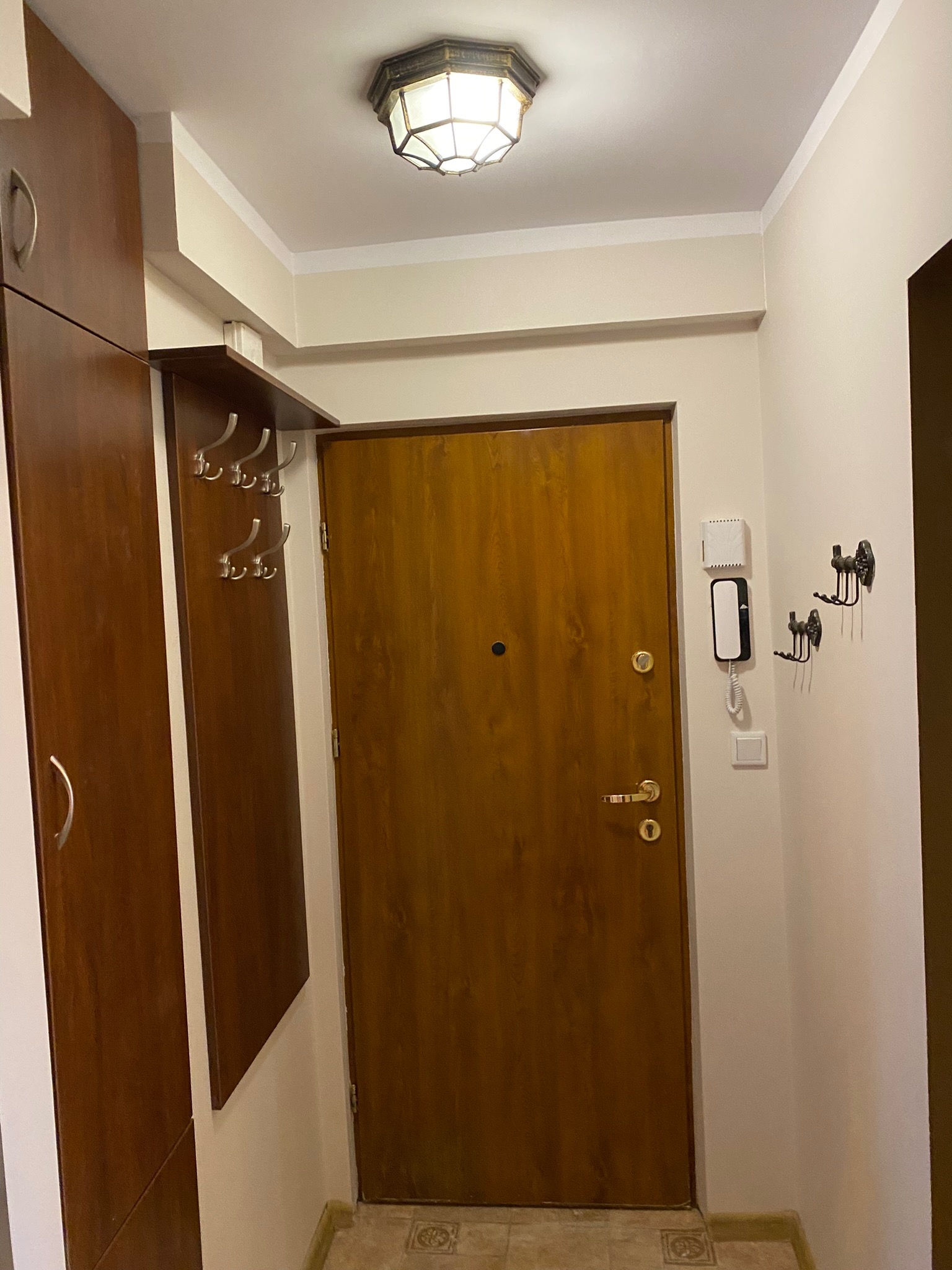 Przedpokój z brązowymi meblami, drewnianymi drzwiami wejściowymi i wieszakami na ścianie, oświetlony lampą sufitową w stylu retro.