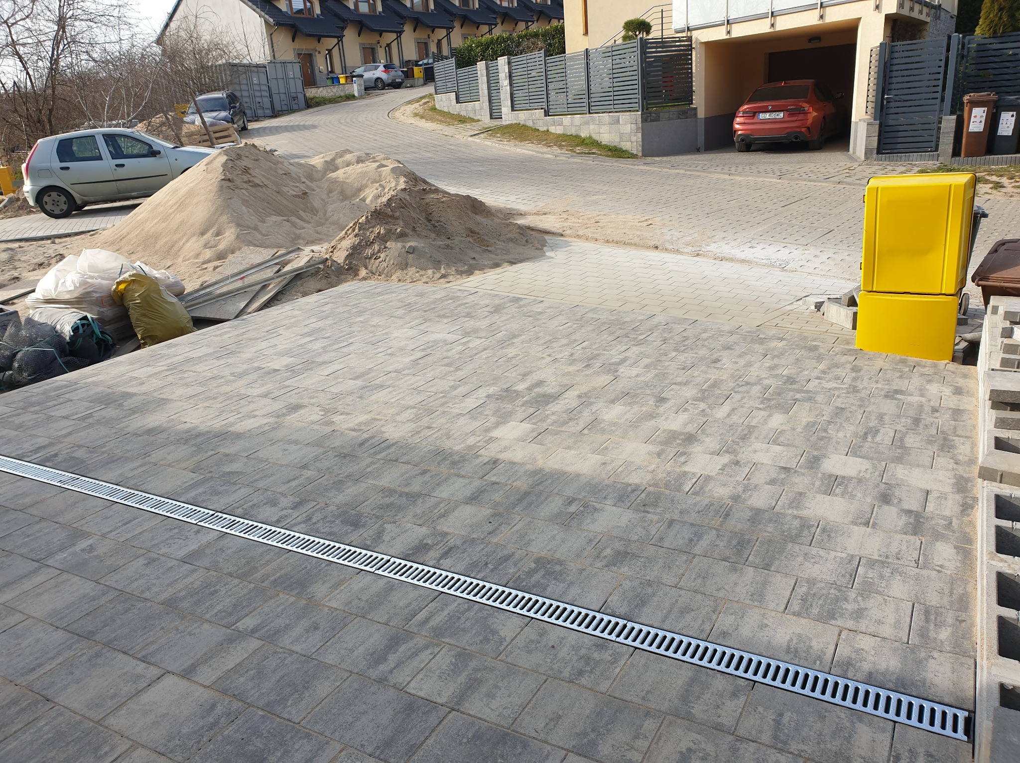 Ułożona szara kostka brukowa z liniowym odpływem wody, w tle hałda piasku, worki z materiałem i zaparkowane samochody.
