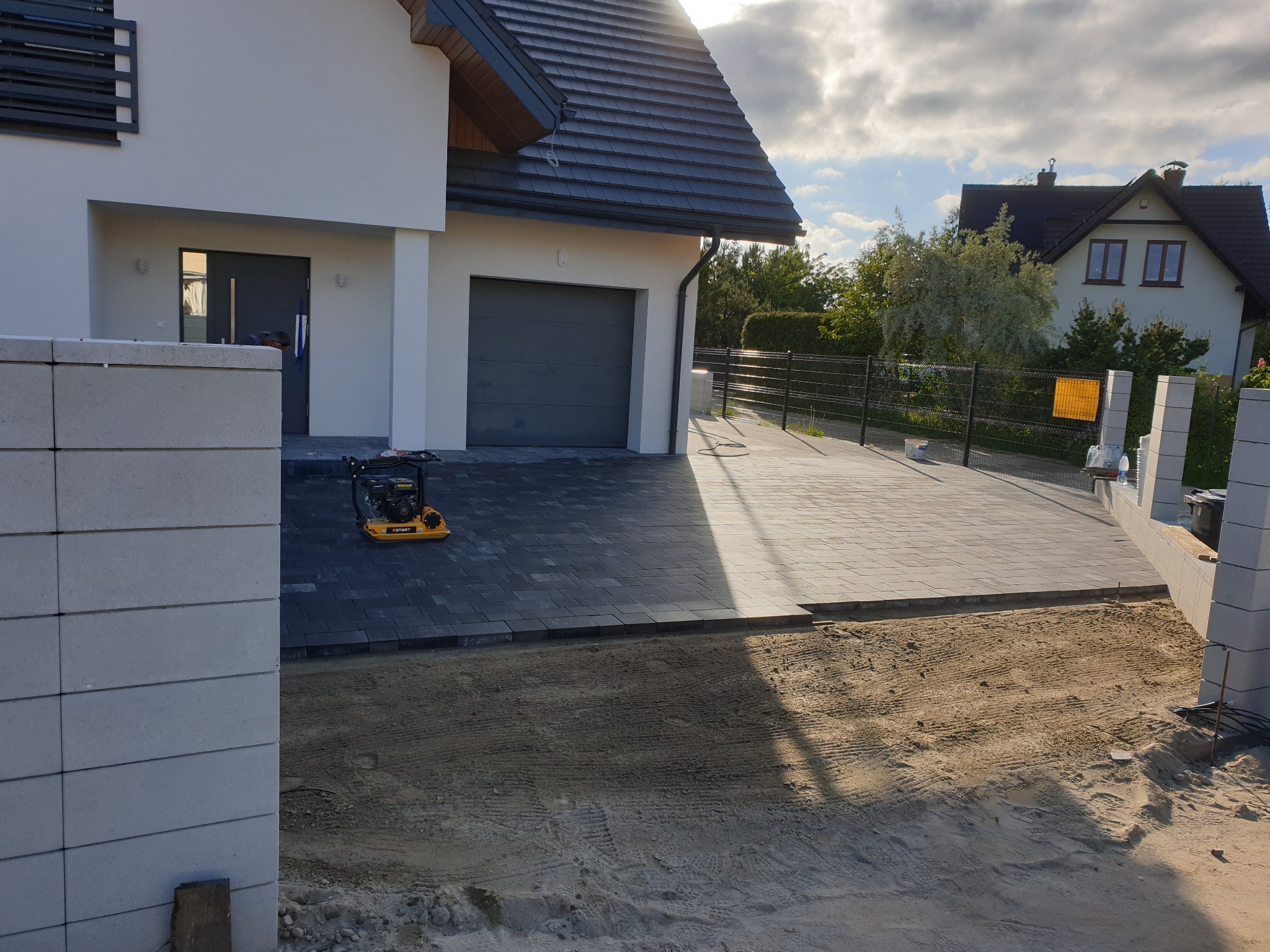 Częściowo ułożony podjazd z szarej kostki brukowej przed nowoczesnym domem jednorodzinnym, widoczna zagęszczarka i przygotowane podłoże z piasku.