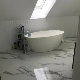 Nowoczesna łazienka na poddaszu z wolnostojącą białą wanną, biało-szarą marmurową podłogą, białą mozaiką na ścianie i oknem dachowym.