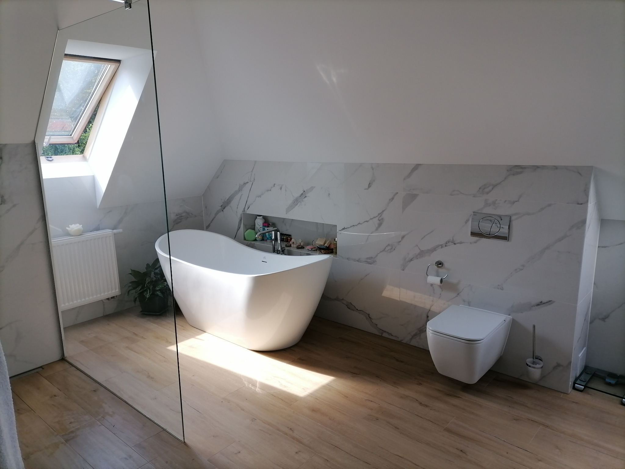 Biała wolnostojąca wanna w nowoczesnej łazience na poddaszu z oknem dachowym, marmurowe płytki ścienne, drewnopodobna podłoga, szklana kabina prysznicowa po lewej stronie.