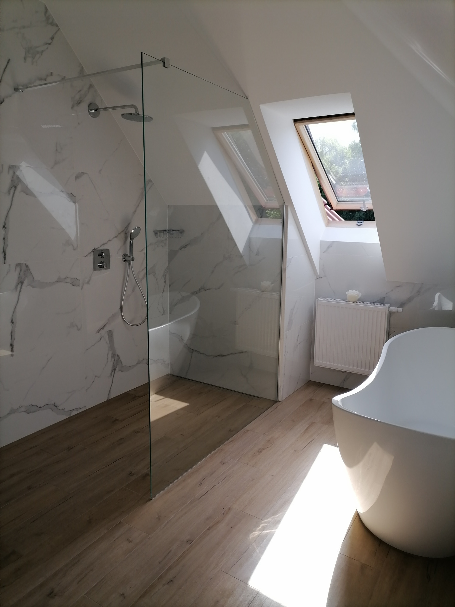 Nowoczesna łazienka na poddaszu z kabiną prysznicową typu walk-in, białą wanną wolnostojącą, marmurowymi płytkami i drewnopodobną podłogą, oświetlona naturalnym światłem z okna dachowego.