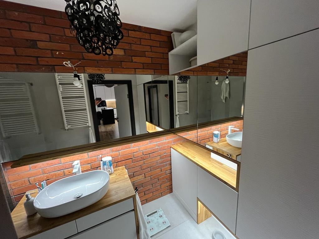 Nowoczesna łazienka z umywalką nablatową na drewnianym blacie, ceglaną ścianą i dużym lustrem z podświetleniem LED, widoczna waga łazienkowa.