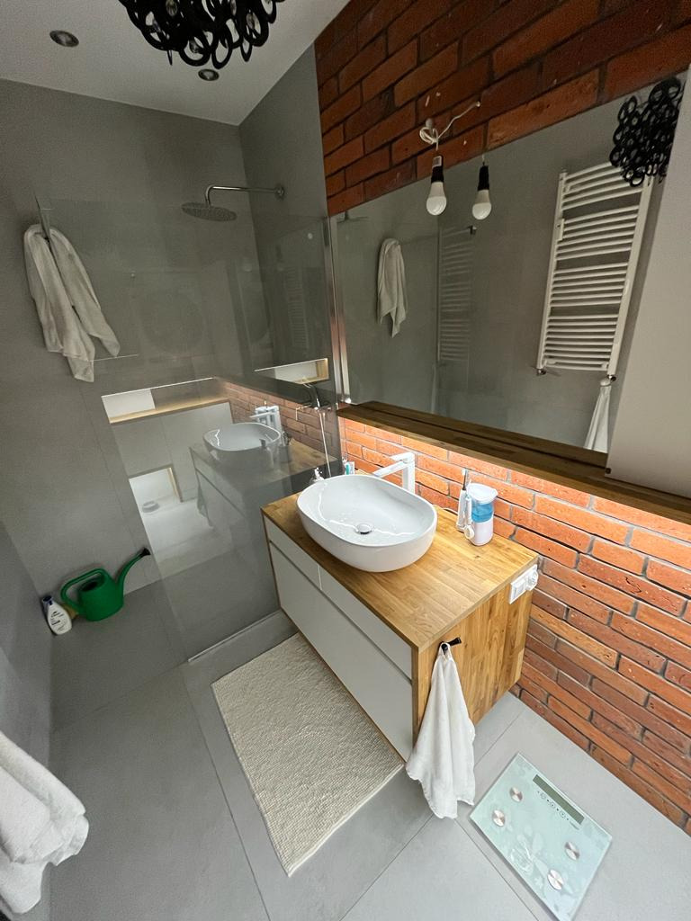 Nowoczesna łazienka z umywalką nablatową na drewnianej szafce, ceglana ściana w tle, widoczne akcesoria łazienkowe i dekoracyjna lampa sufitowa.