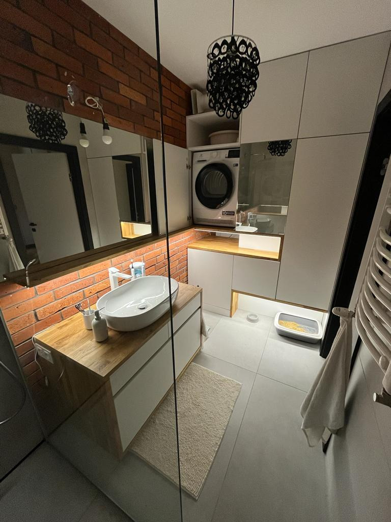 Nowoczesna łazienka z ceglaną ścianą, owalną umywalką na drewnianej szafce, pralką w zabudowie i dekoracyjną lampą wiszącą.