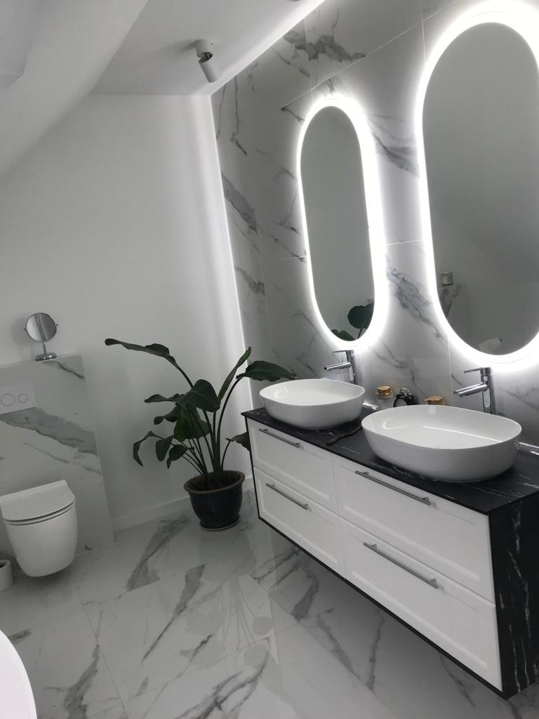 Nowoczesna łazienka w bieli i szarości z marmurowym wzorem, podwójna umywalka nablatowa na białej szafce z czarnym blatem, podświetlane lustra owalne i roślina doniczkowa.