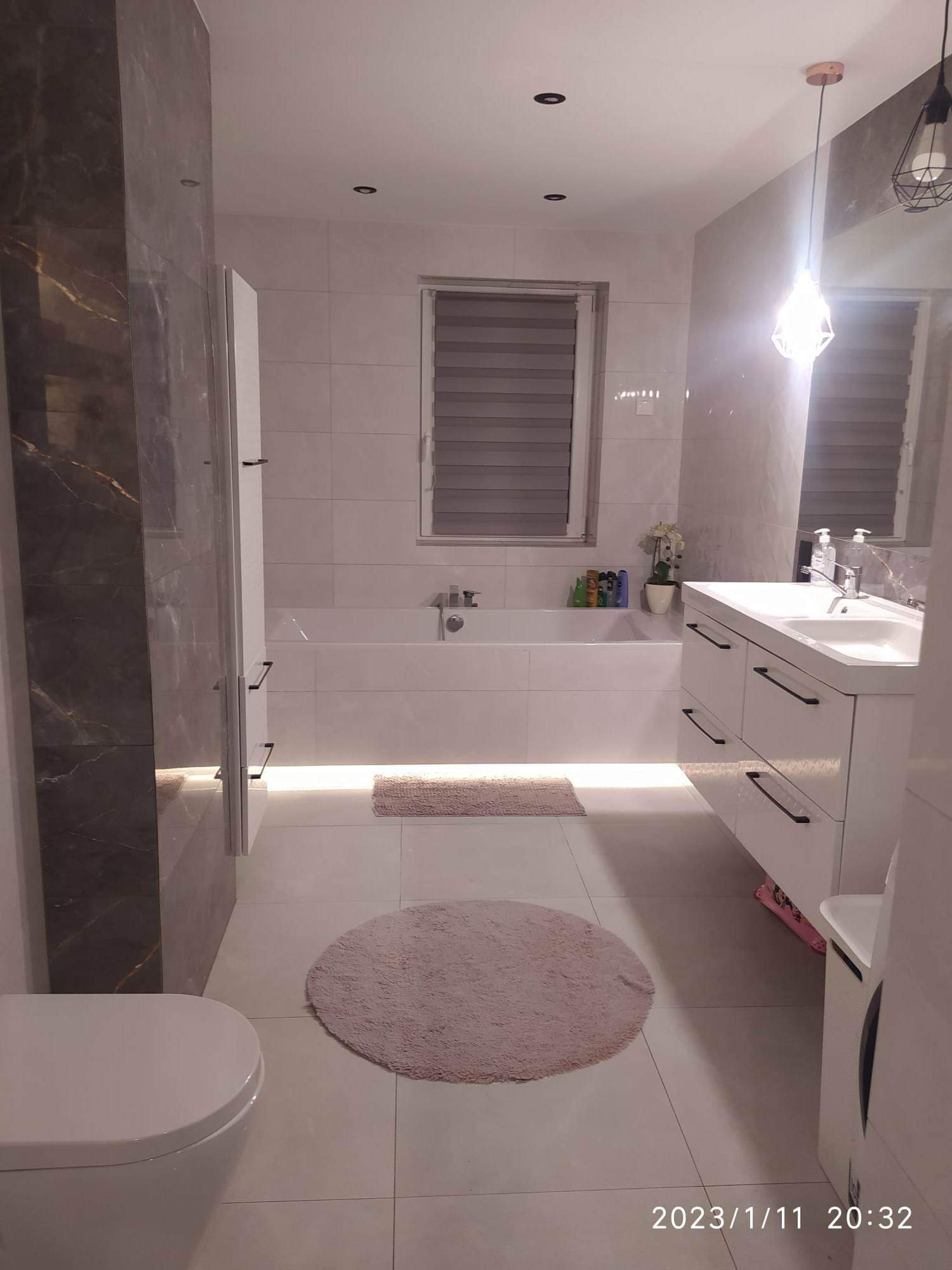 Nowoczesna łazienka z białymi płytkami, podświetlaną wanną, umywalką z szafką i ciemnoszarą ścianą z marmurowym wzorem przy kabinie prysznicowej.