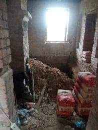 Wnętrze łazienki w trakcie remontu: surowe ściany z cegły, stos worków z zaprawą murarską, widoczne narzędzia budowlane, okno z silnym światłem dziennym. W tle ściana z widoczną dziurą po instalacji.