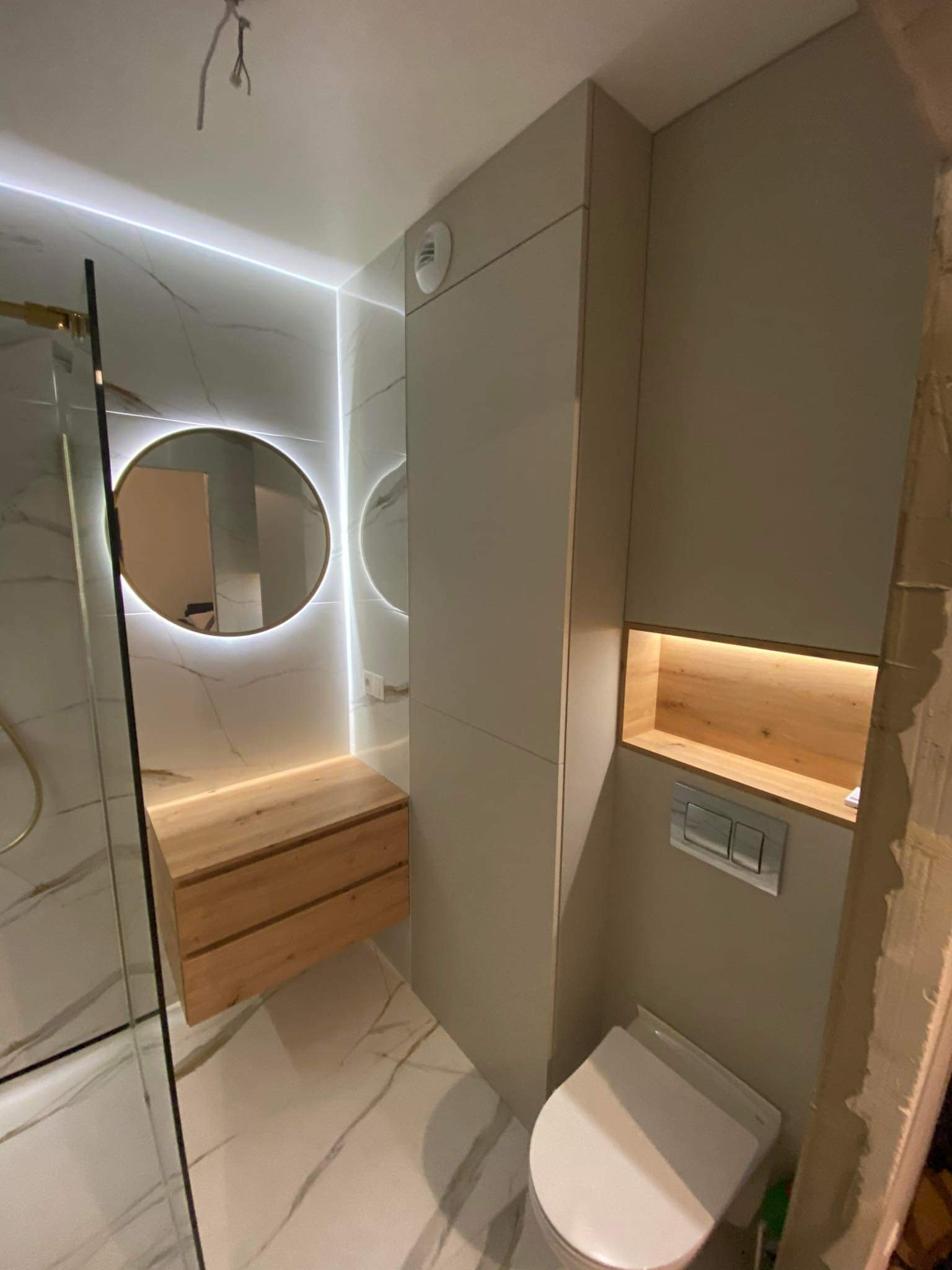 Nowoczesna łazienka z podświetlanym lustrem, drewnianą szafką pod umywalką i marmurowymi płytkami, widoczne kable wystające z sufitu.