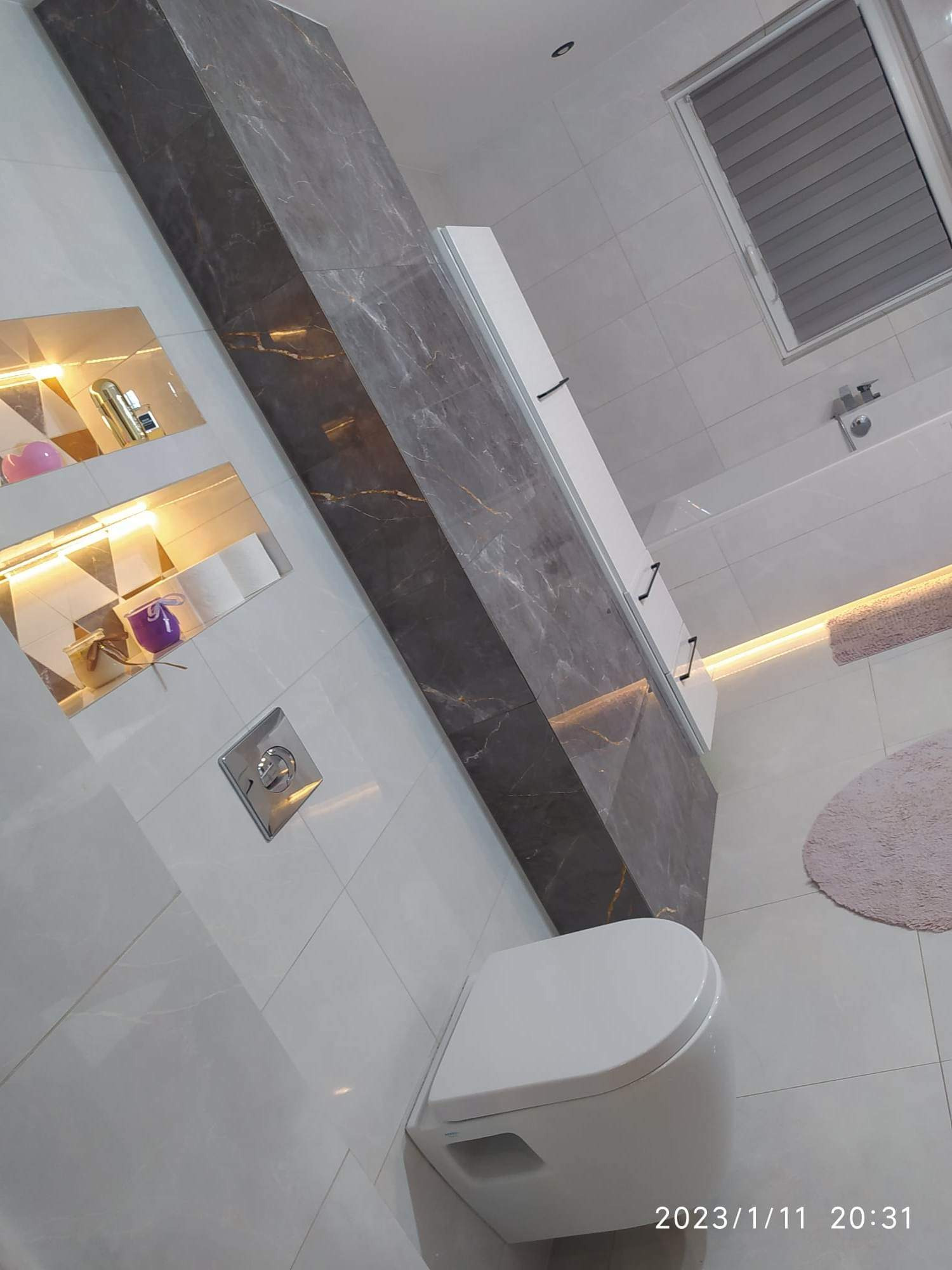 Nowoczesna łazienka z podświetlanymi półkami, marmurową ścianą akcentującą, wiszącą toaletą i szarą roletą w oknie.