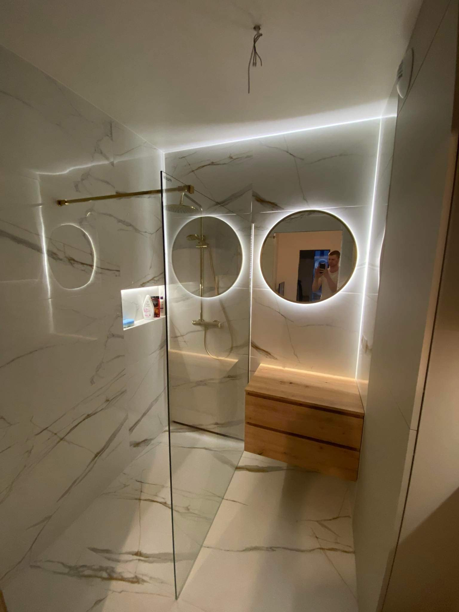 Nowoczesna łazienka z marmurowymi płytkami, kabiną prysznicową ze złotymi elementami i okrągłymi lustrami z podświetleniem LED, drewniana szafka pod umywalką.