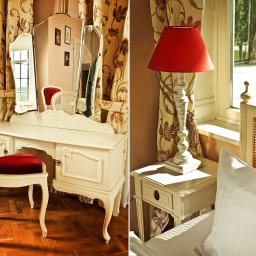 Studio Migafka - Jasna toaletka z trzema lustrami, krzesłem z czerwonym siedziskiem i drewnianą podłogą w pokoju z zasłonami w kwiaty oraz stolik nocny z lampką z czerwonym abażurem