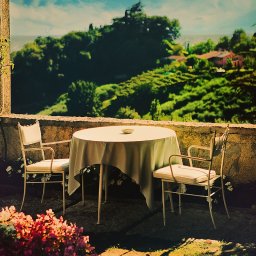 Studio Migafka - Ustawiony stół z dwoma krzesłami na tarasie z widokiem na toskański krajobraz, zarośnięte wzgórza i domy w oddali, ujęcie z niskiego kąta przez roślinność, ciepłe światło.