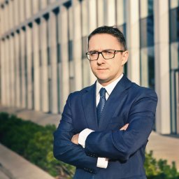 Studio Migafka - Portret mężczyzny w okularach i granatowym garniturze, z założonymi rękami na tle nowoczesnego biurowca w słoneczny dzień.