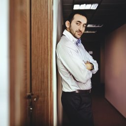 Studio Migafka - Mężczyzna w koszuli w paski opiera się o ścianę w korytarzu biurowym, z założonymi rękami i zegarkiem na nadgarstku.