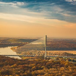 Studio Migafka - Wysoka konstrukcja mostu wiszącego nad rzeką, widok z góry na jesienny krajobraz z lasami i polami w ciepłym świetle zachodzącego słońca.