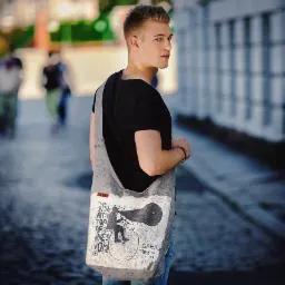 Młody mężczyzna z blond włosami, ubrany w czarny t-shirt i jeansy, patrzy przez ramię. Na ramieniu ma szarą torbę z nadrukiem graficznym przedstawiającym postać na rowerze z megafonem. W tle...